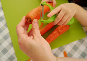 Małe rączki dziecka, pod opieką nauczyciela, próbują obierać marchewkę.